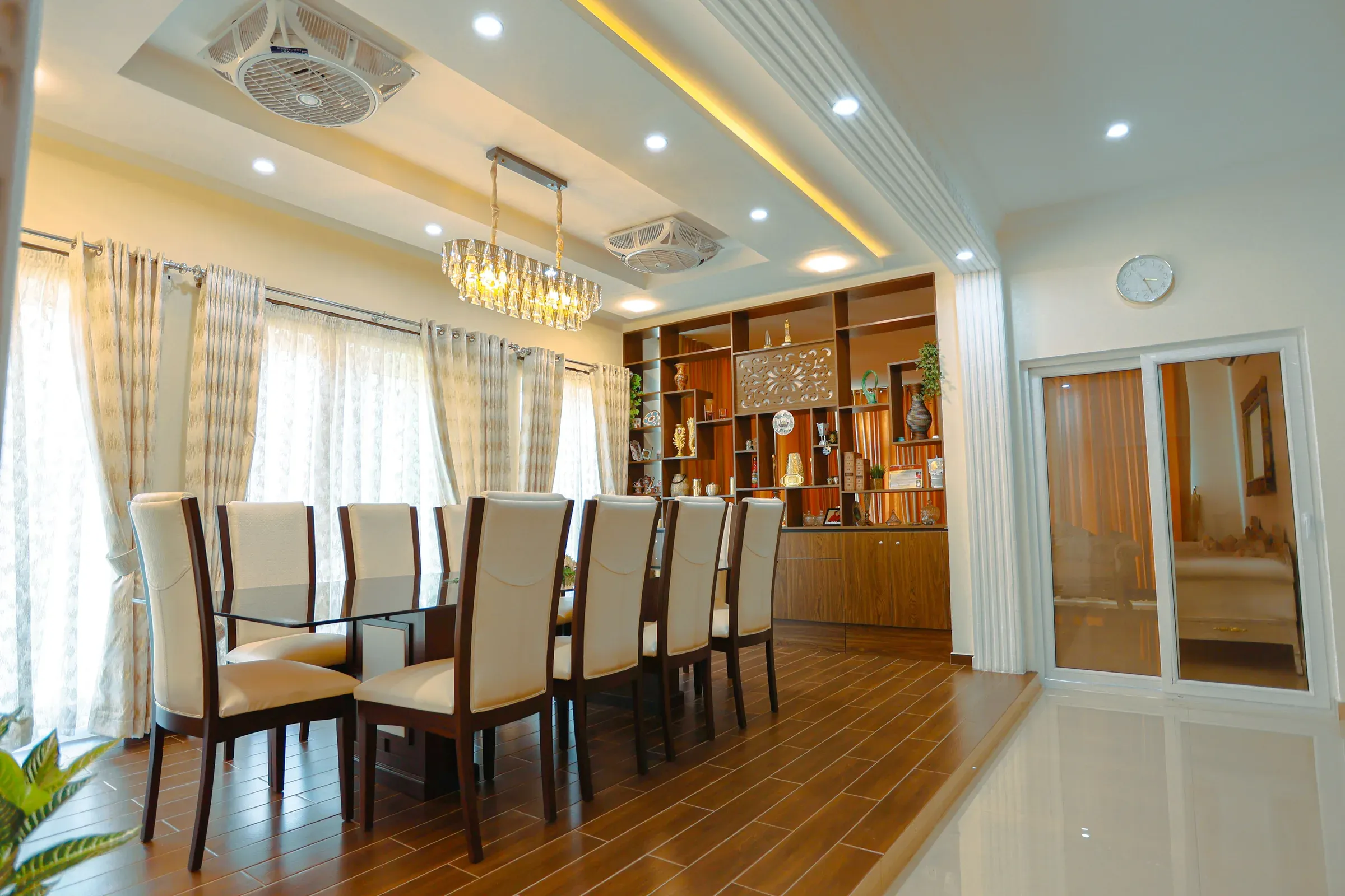 Dining room with a crystal chandelier, eight beige chairs, a glass table and a wooden display unit