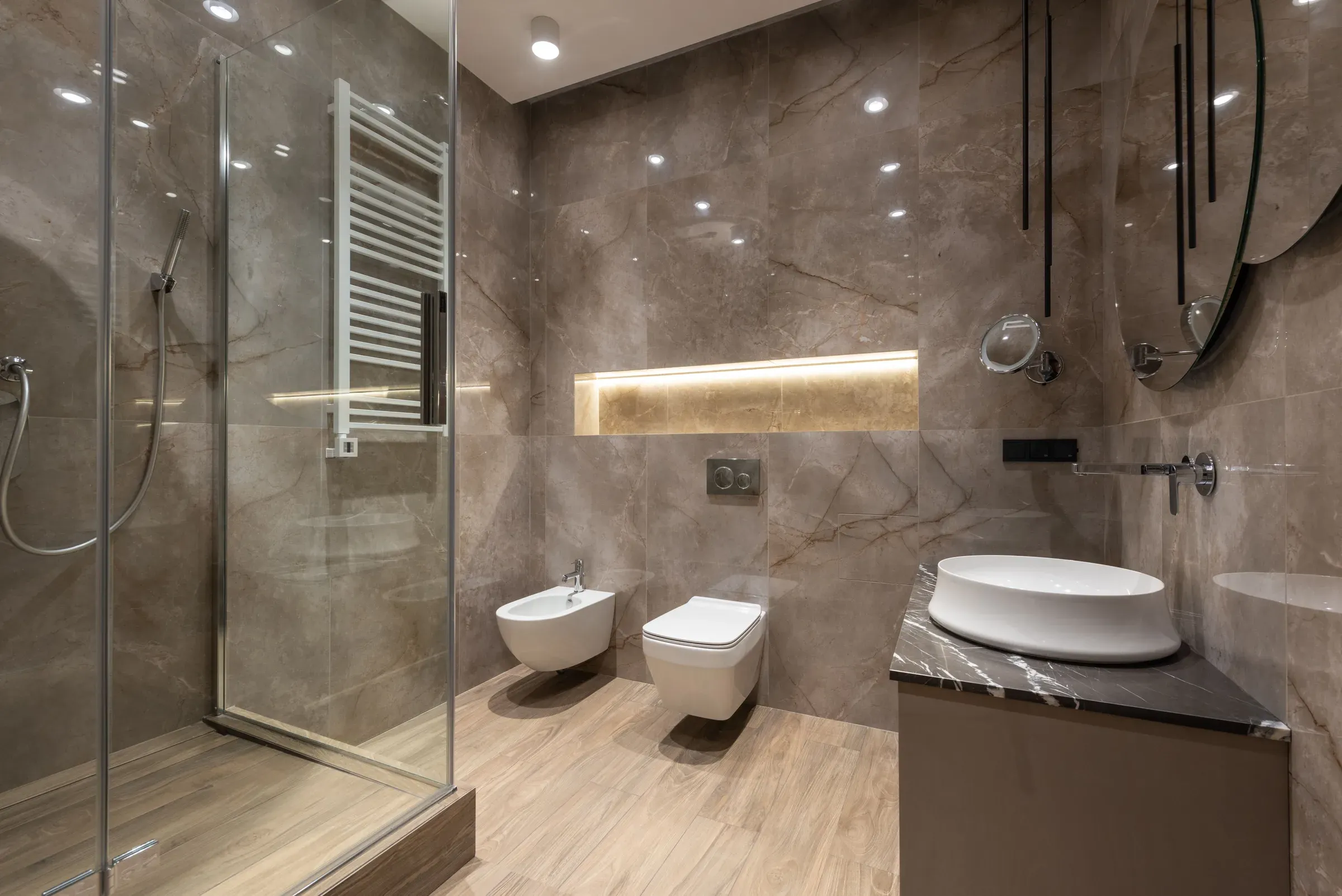 Beige marble-tile bathroom with a glass shower, wall-hung WC, lit wall niche and a vessel basin on a dark marble counter
