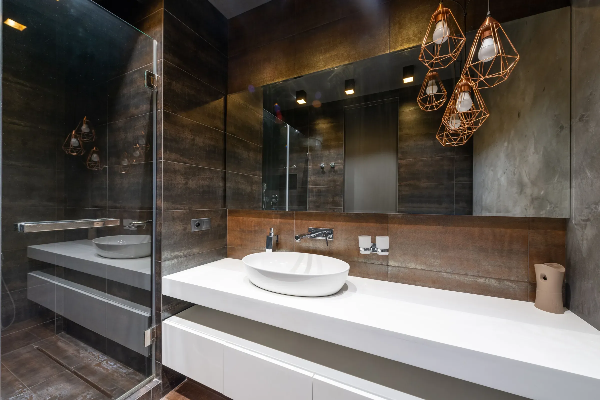 Dark wood-look tile bathroom with a white floating counter, vessel basin and copper geometric pendant lights