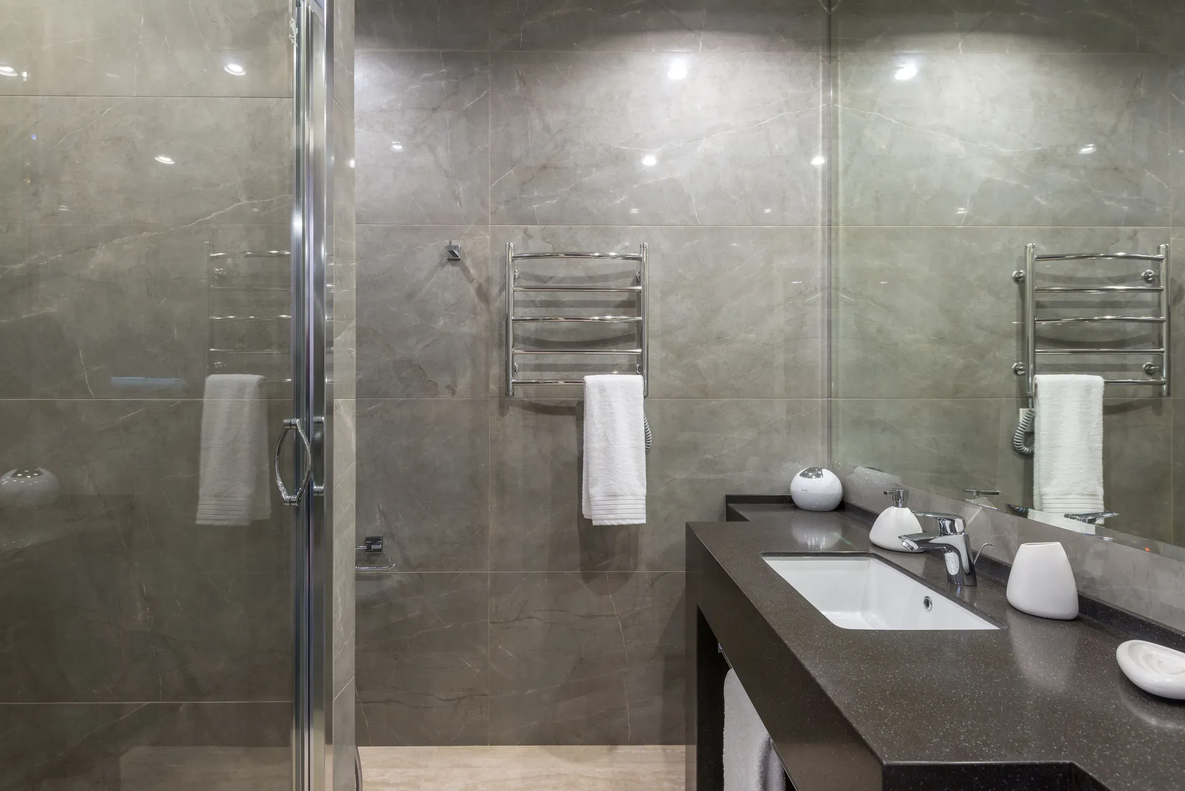 Grey large-format tile bathroom with a dark quartz vanity, undermount basin, towel warmer and glass shower