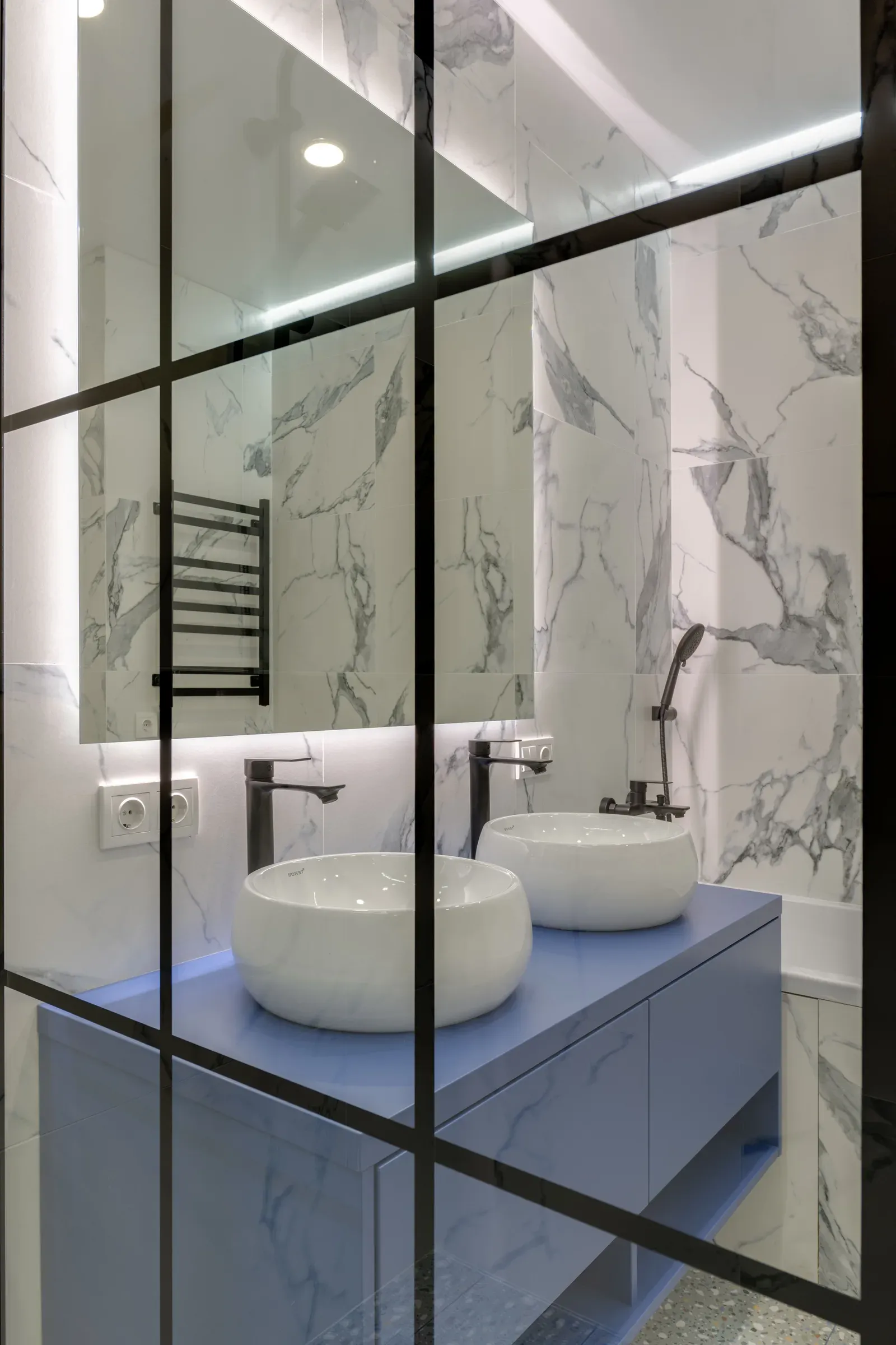 White marble-look bathroom behind a black-framed glass partition, with a blue vanity, two round vessel basins and a backlit mirror