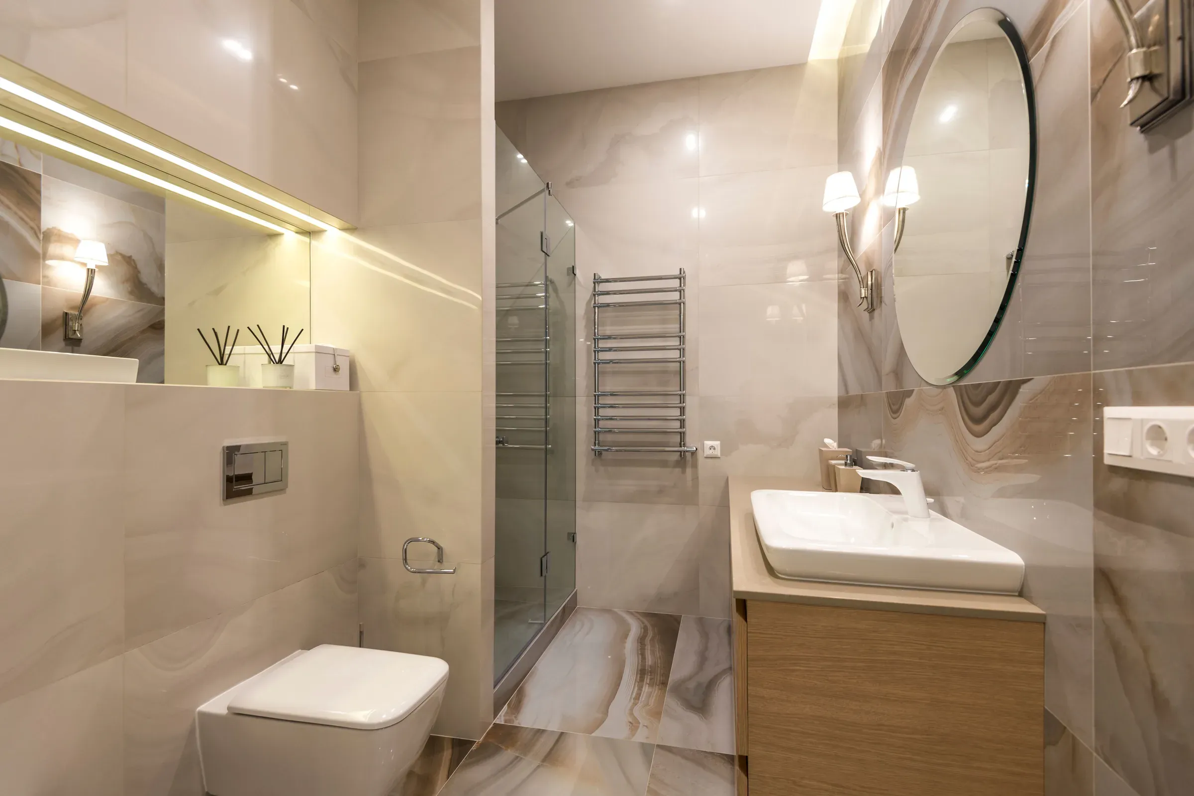 Beige onyx-look tiled bathroom with a wooden vanity, vessel basin, round mirror with sconces and glass shower