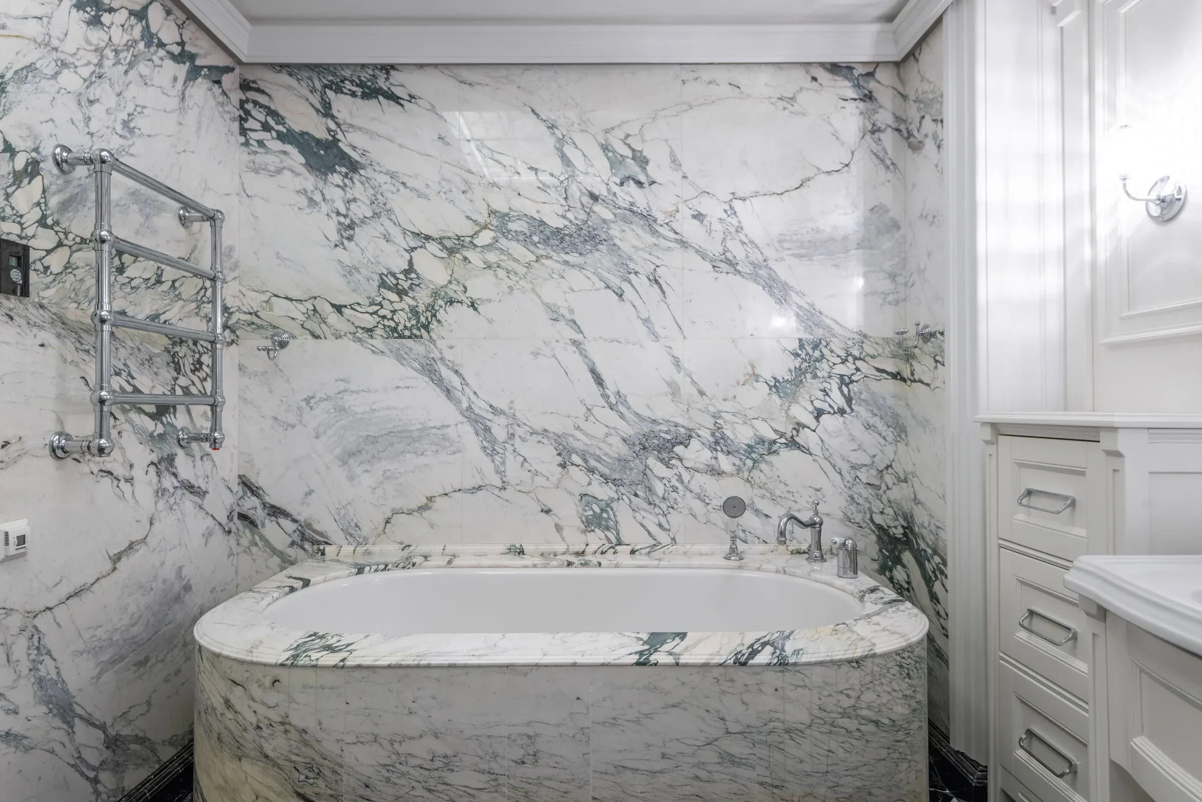 Bathroom clad in white marble with bold grey-green veins, a marble-faced bathtub and white panelled cabinets