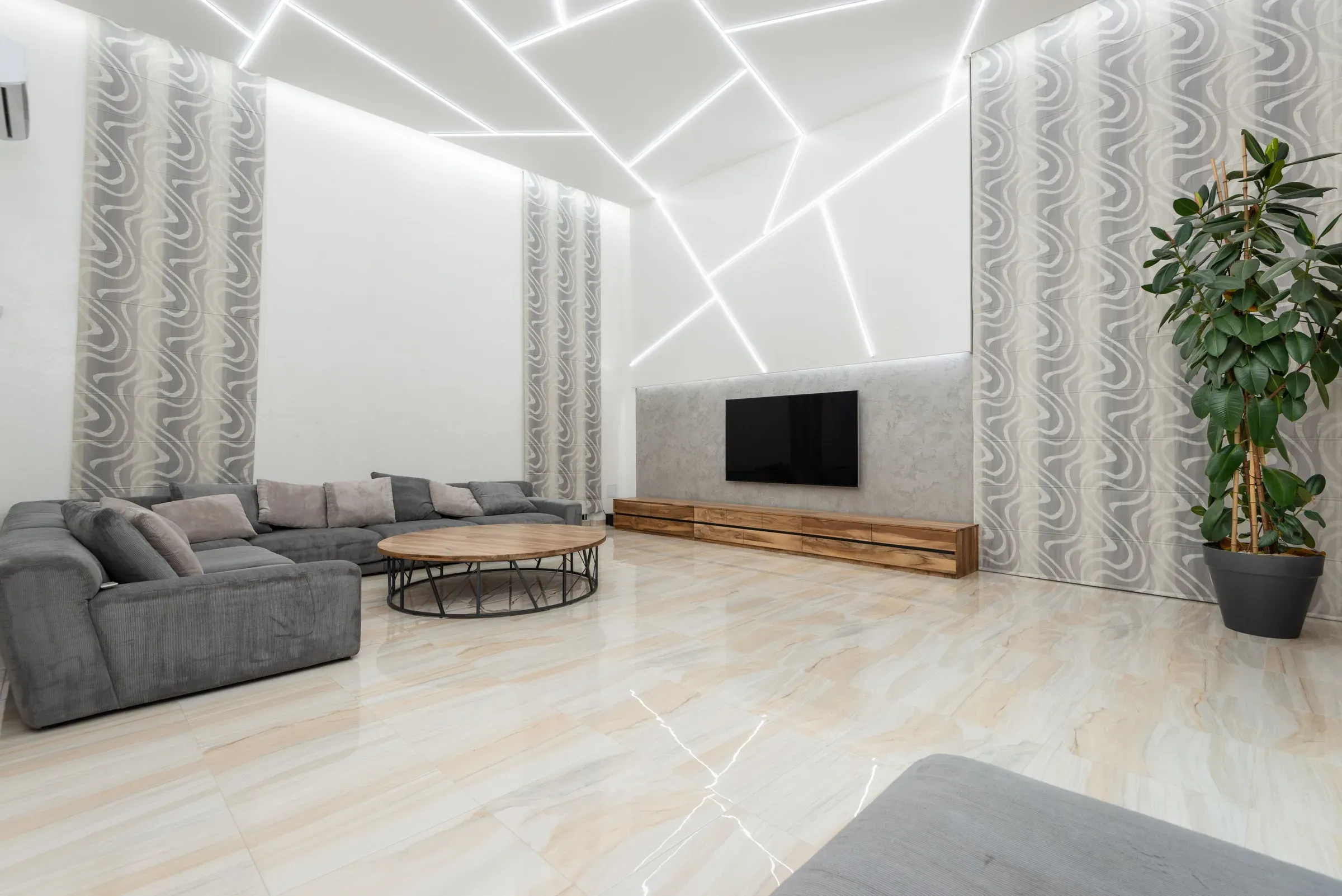 Living room with a geometric LED-line false ceiling, grey sectional sofa, TV on a concrete wall and marble-look floor