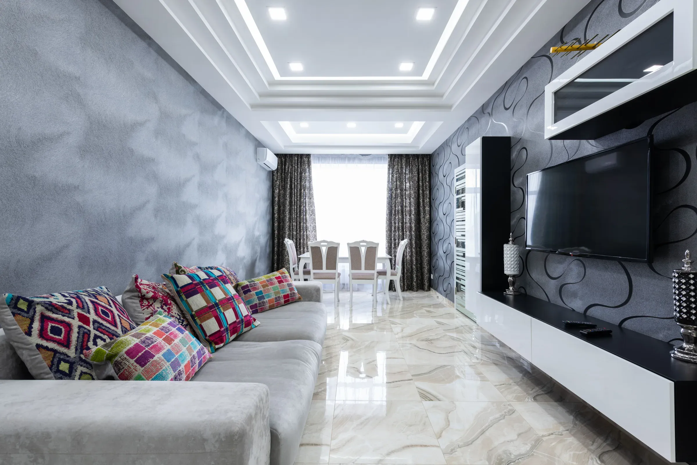 Long living room with a layered POP ceiling, grey sofa with colourful cushions and dining area by the window