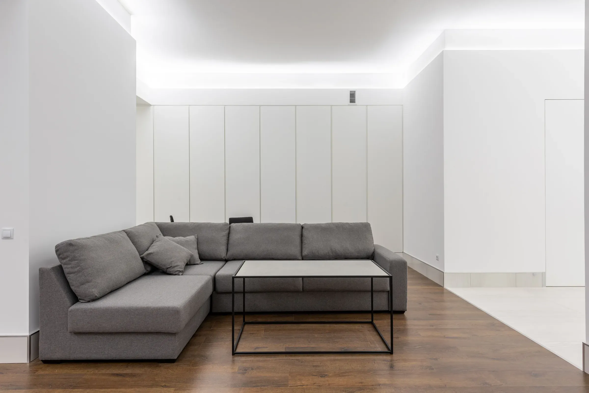 Minimal white living room with perimeter cove lighting, handleless white wall cabinets and a grey L-shaped sofa