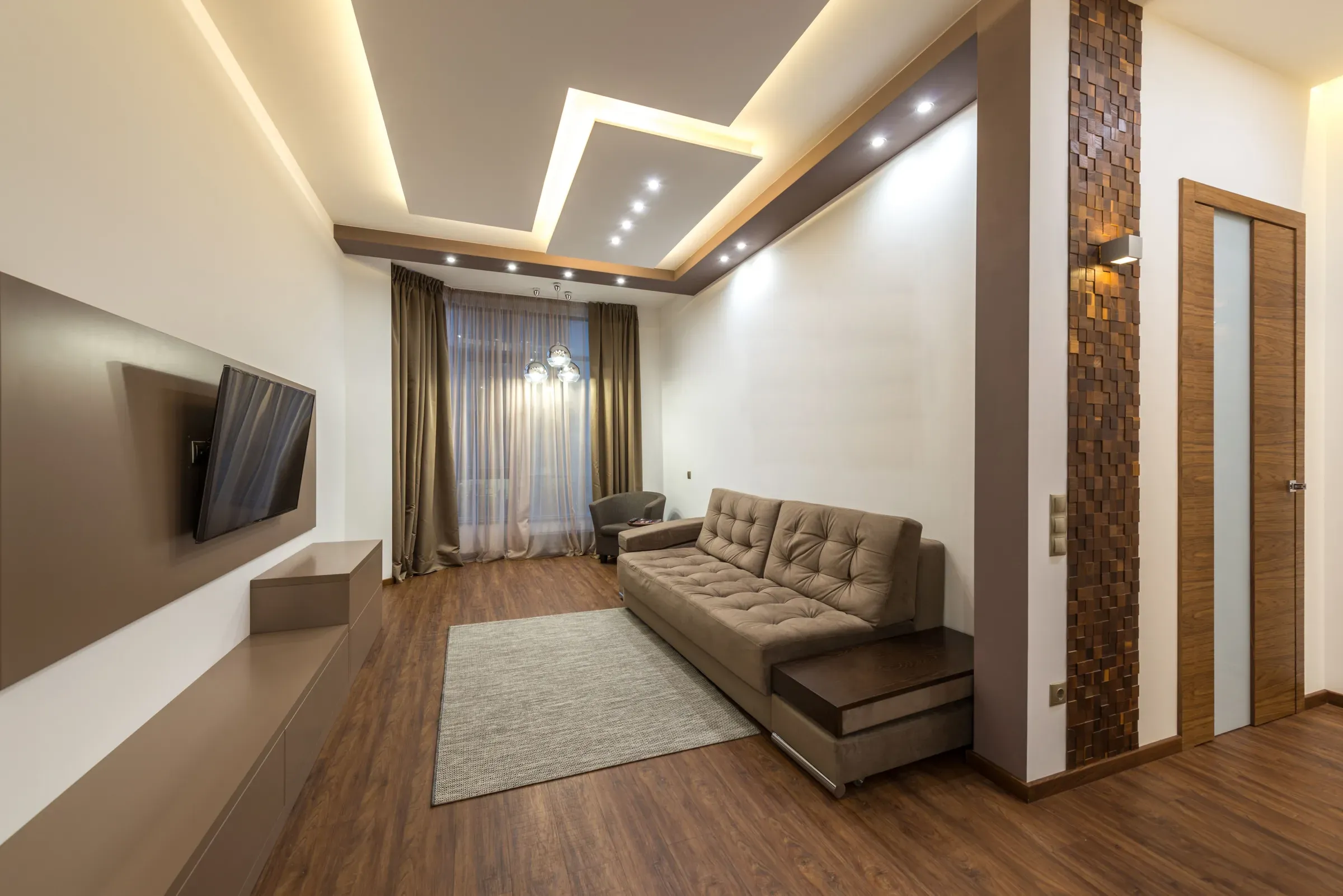 Living room with a stepped cove-lit ceiling, brown fascia with spotlights, beige sofa and wooden mosaic pillar