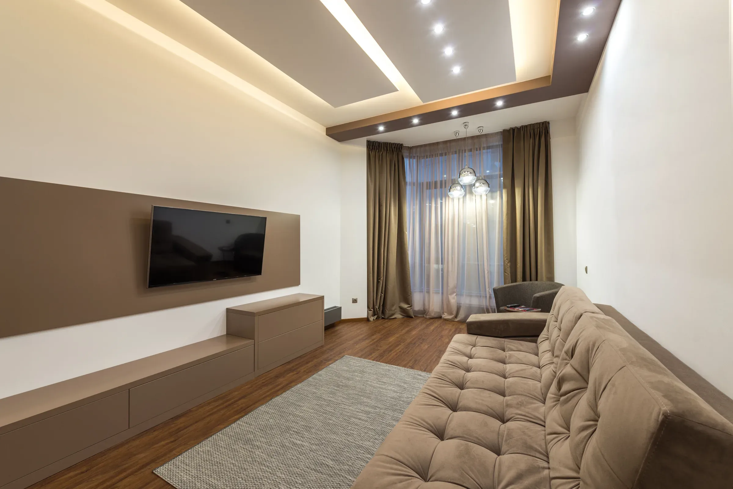 Living room with a two-level false ceiling, cove light and spotlights, tufted sofa and TV on a brown wall panel