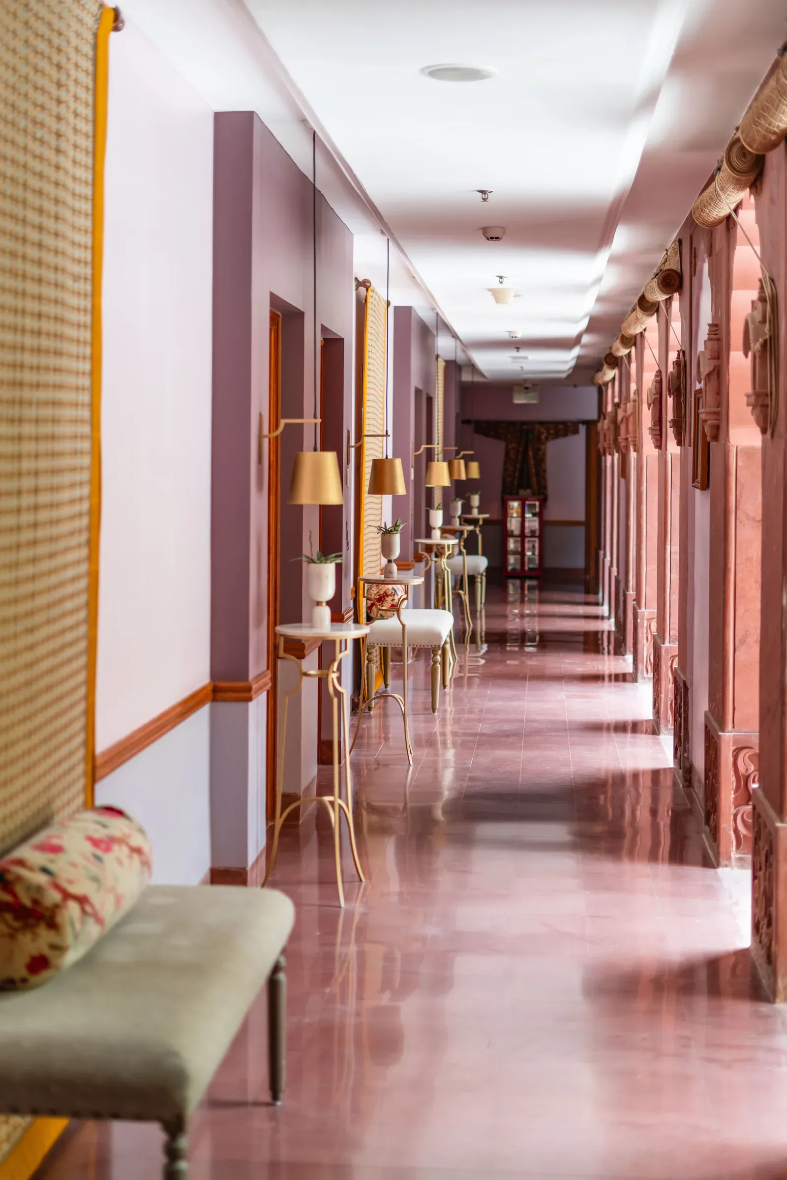 Long heritage corridor with a pink stone floor, carved arches, brass floor lamps and an upholstered bench