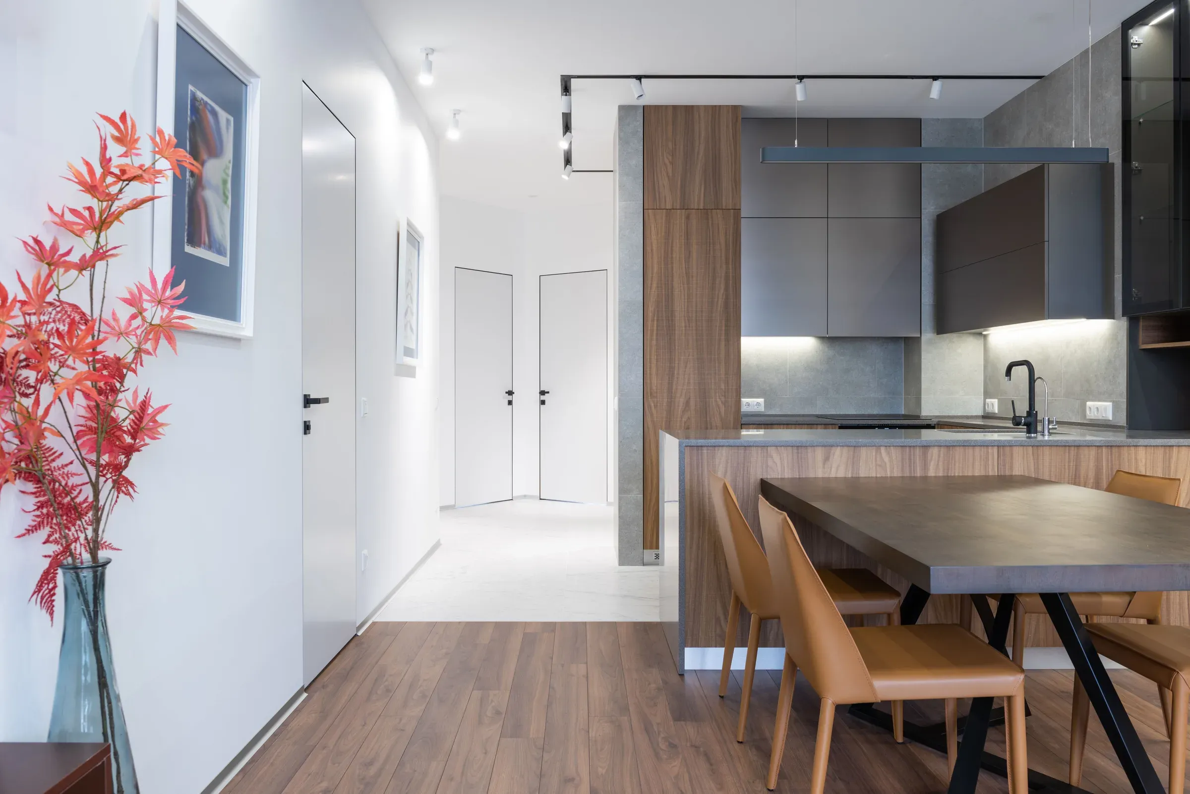 Open kitchen with a walnut tall unit, grey handleless cabinets, track lights and a dining table with tan chairs