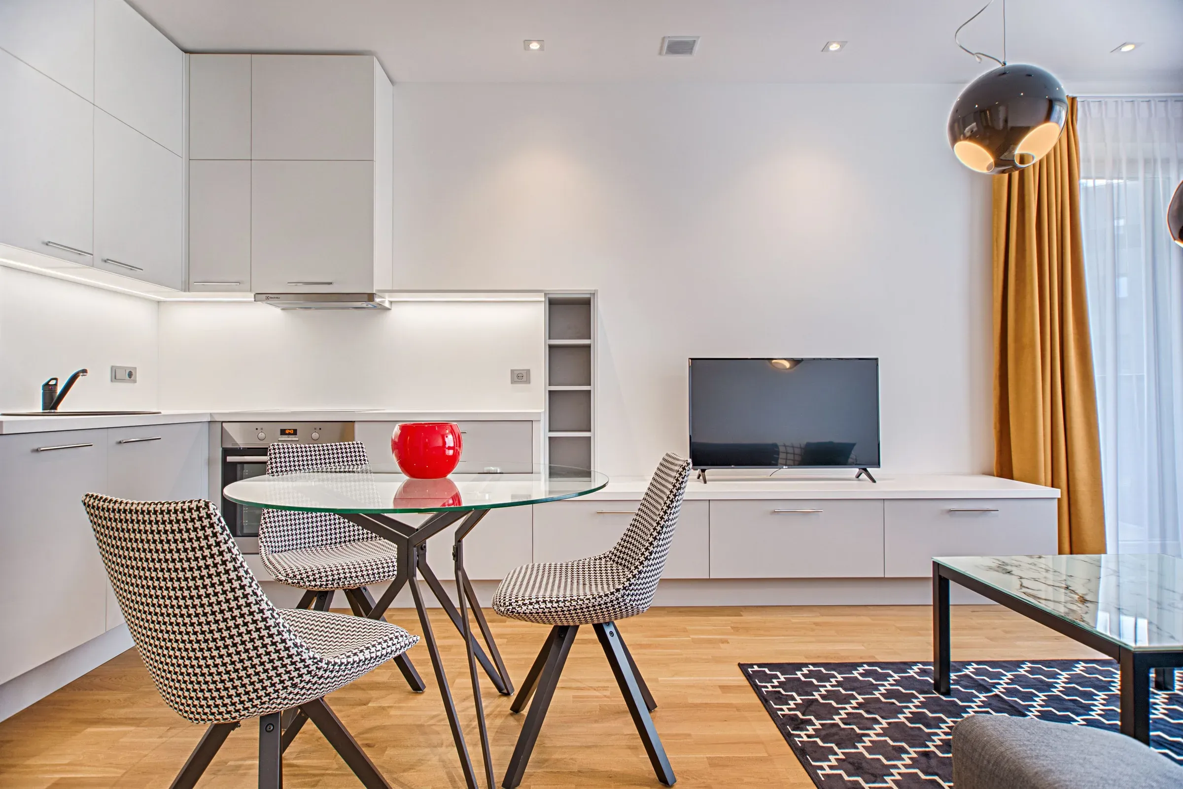 Compact white kitchen with a round glass dining table, houndstooth chairs and a TV unit beside it