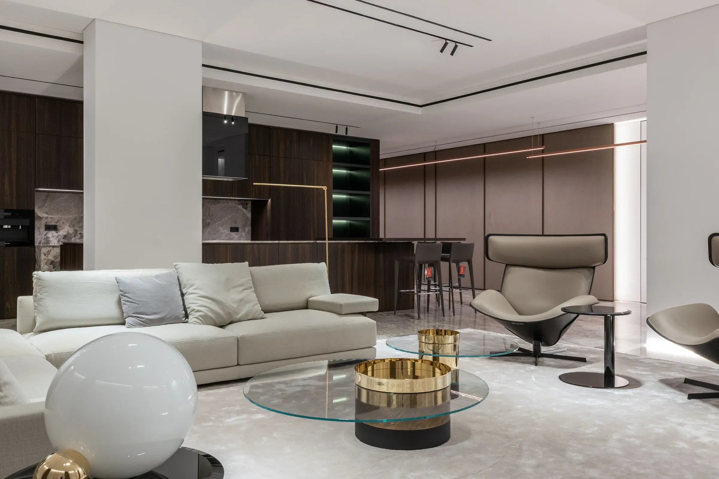 Contemporary living room with a beige sofa, glass and brass coffee tables, lounge chair and open walnut kitchen