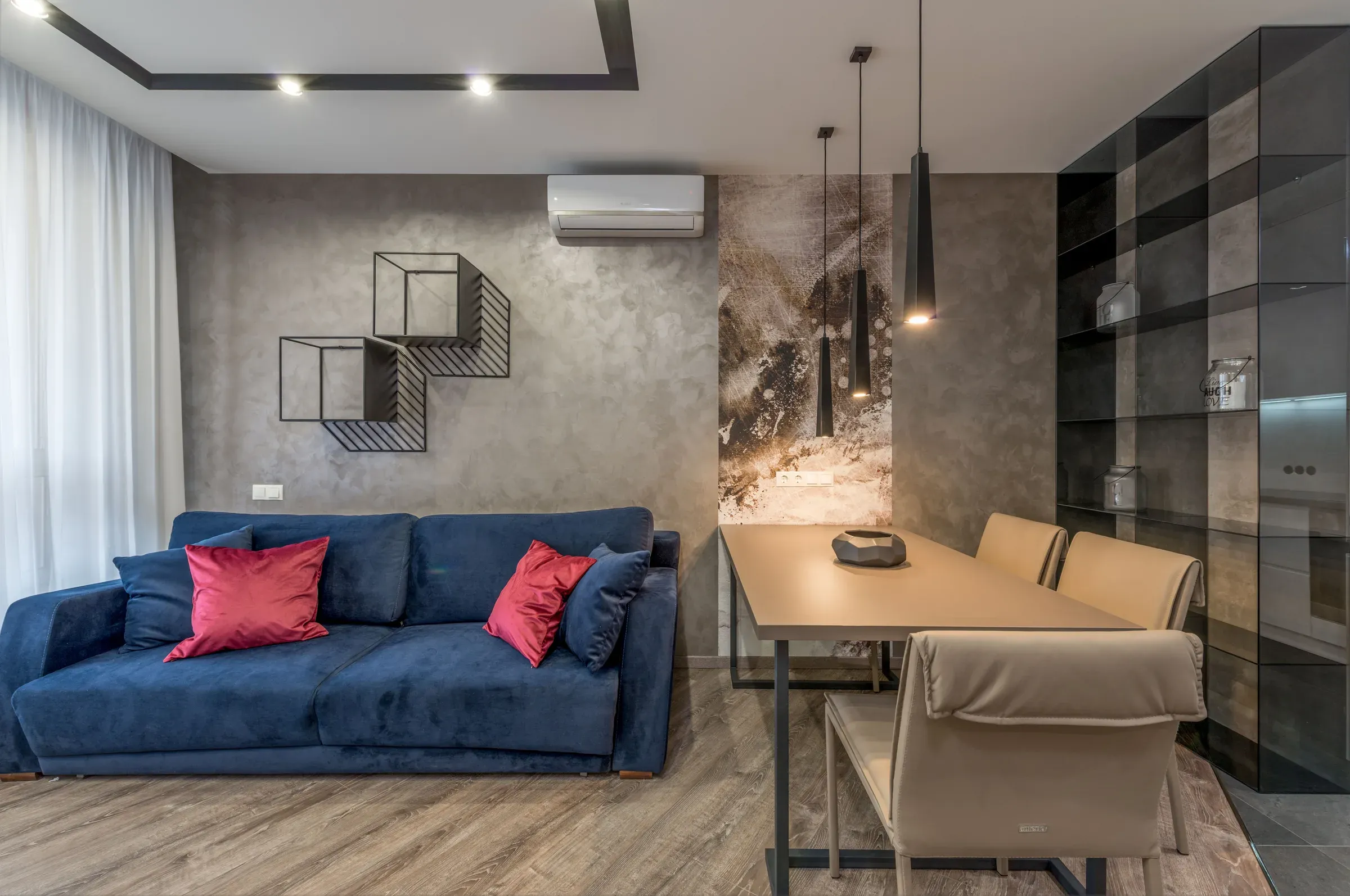 Living-dining room with a blue velvet sofa, metal cube wall shelves, grey textured wall and wooden dining table