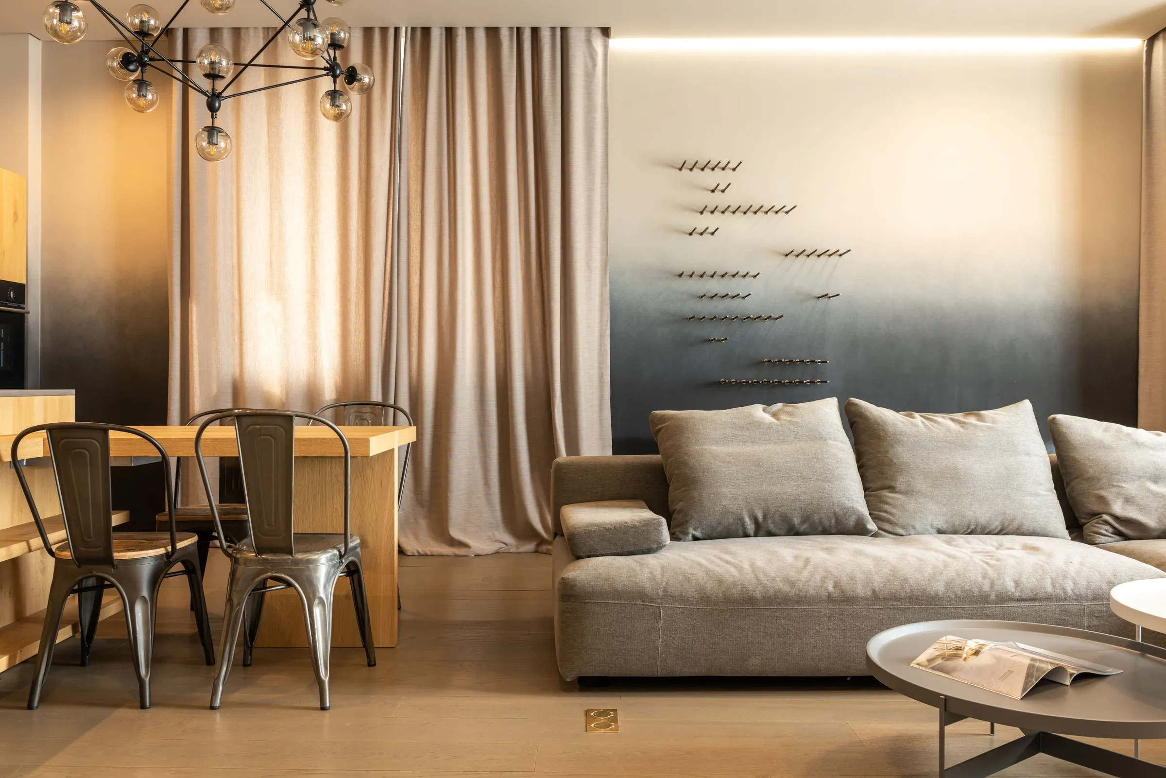 Living room with a grey sofa, ombre wall with metal wall sculpture, wooden dining table and metal chairs