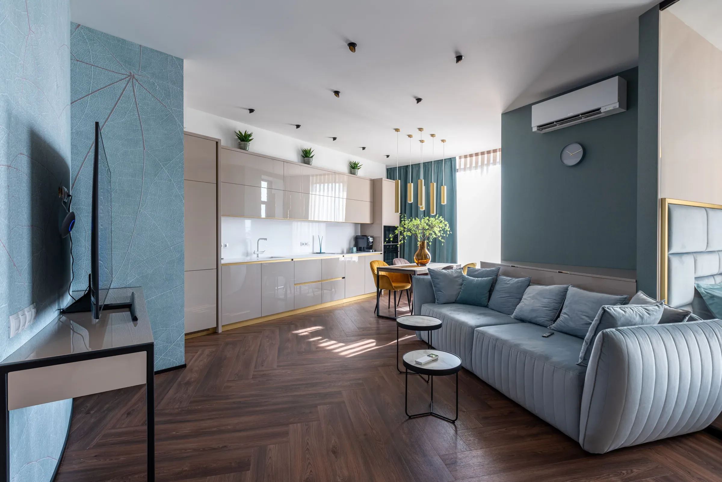 Open living room with a herringbone wooden floor, grey channel sofa, teal walls and a beige kitchen
