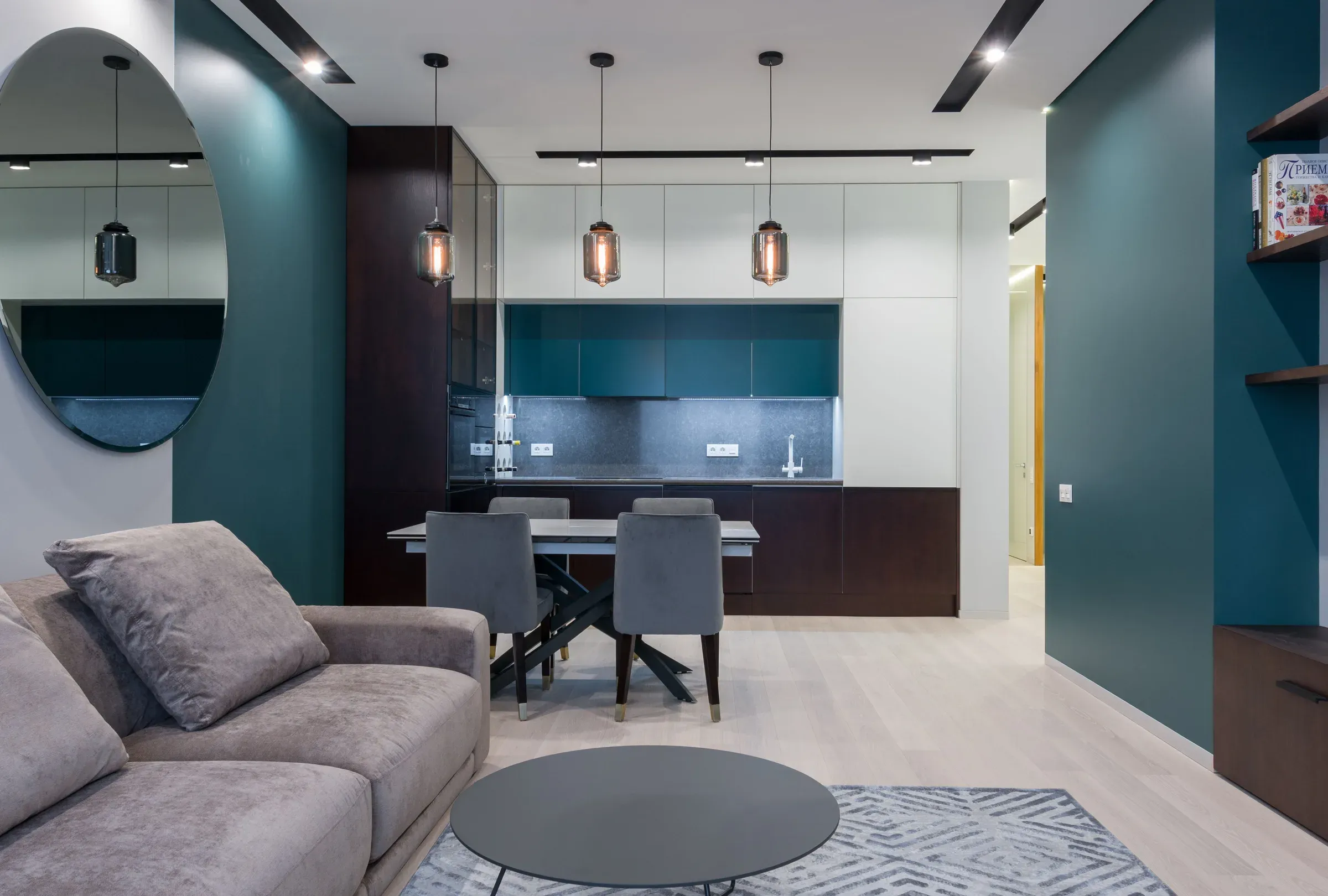 Living-dining room with teal walls, a round mirror, grey sofa and a kitchen with pendant lights