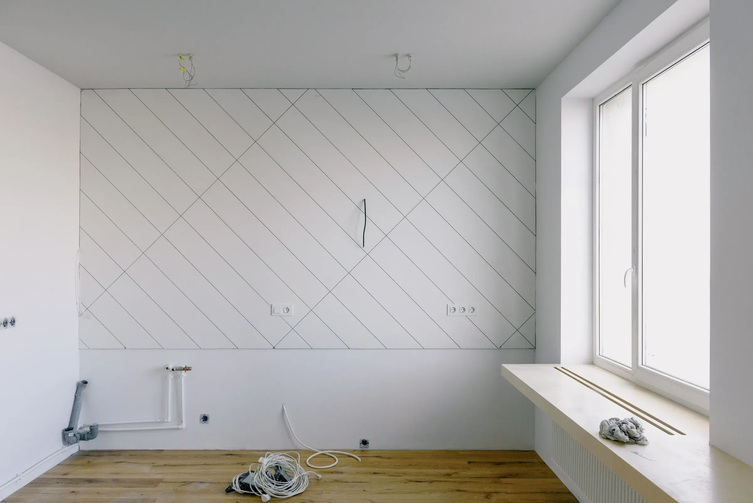 Room under renovation with a bare white wall marked for diagonal tiles, exposed pipes and a new wooden floor