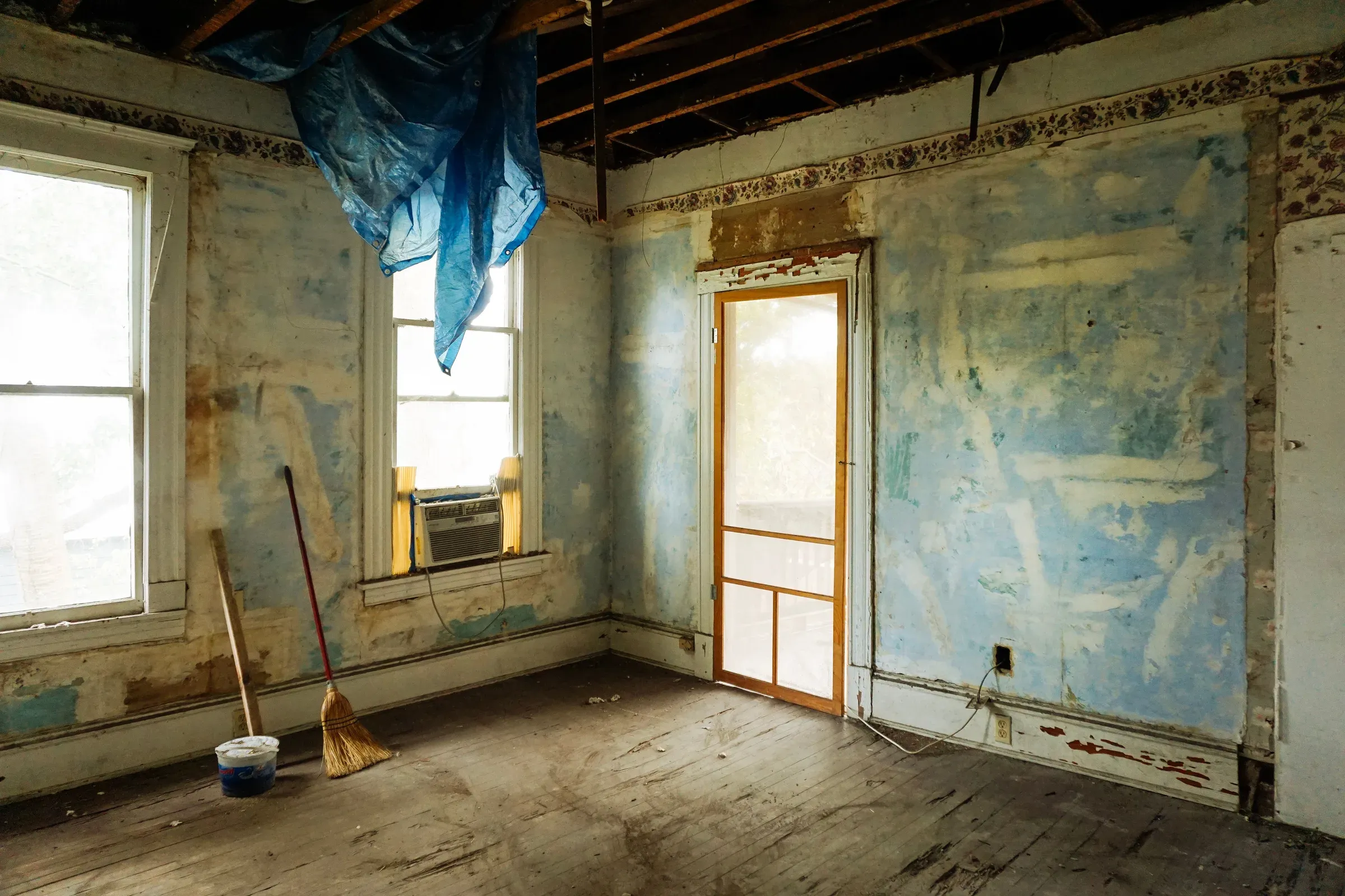 Old room before renovation with peeling blue paint, exposed ceiling joists, worn floorboards and a broom