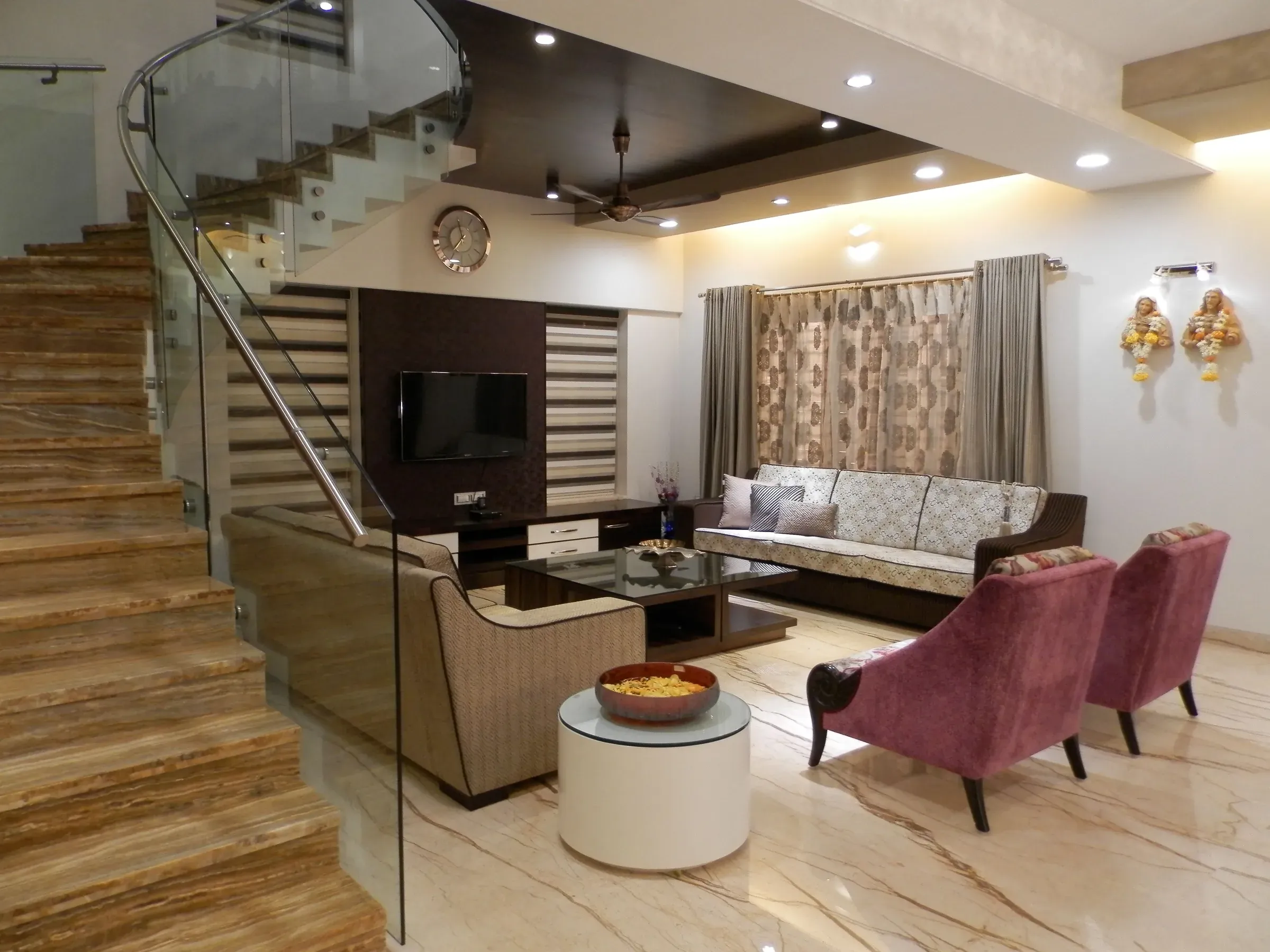 Indian duplex living room with a marble-look staircase, glass railing, TV unit and pink armchairs