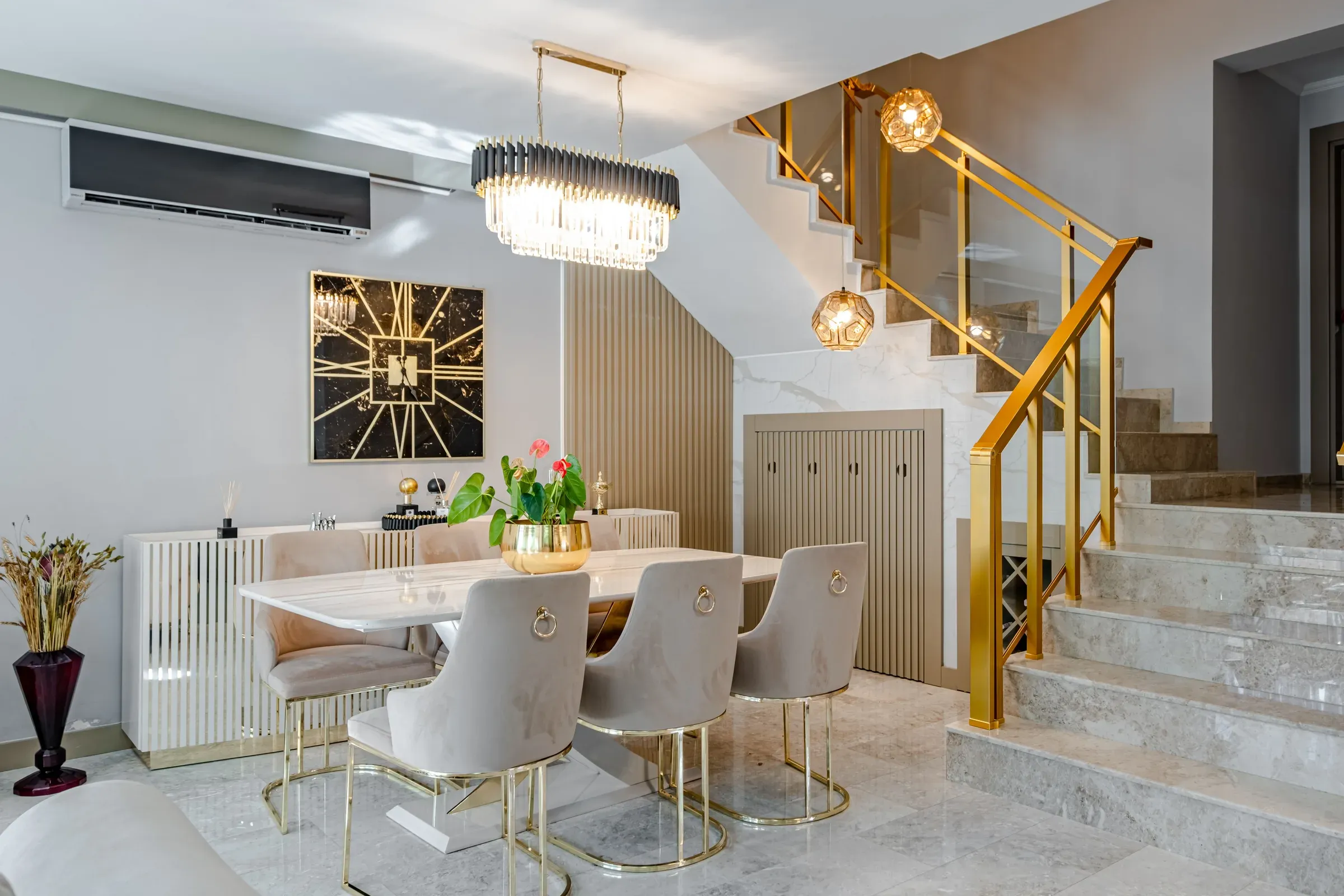 Dining area beside a marble staircase with a gold glass railing, crystal chandelier and fluted wall panel