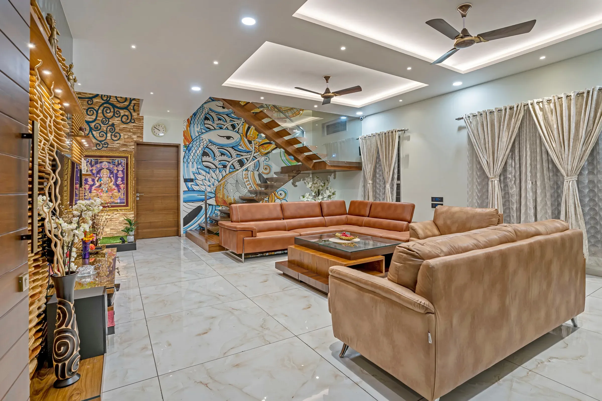 Indian duplex living room with a painted mural on the staircase wall, tan leather sofas and ceiling fans