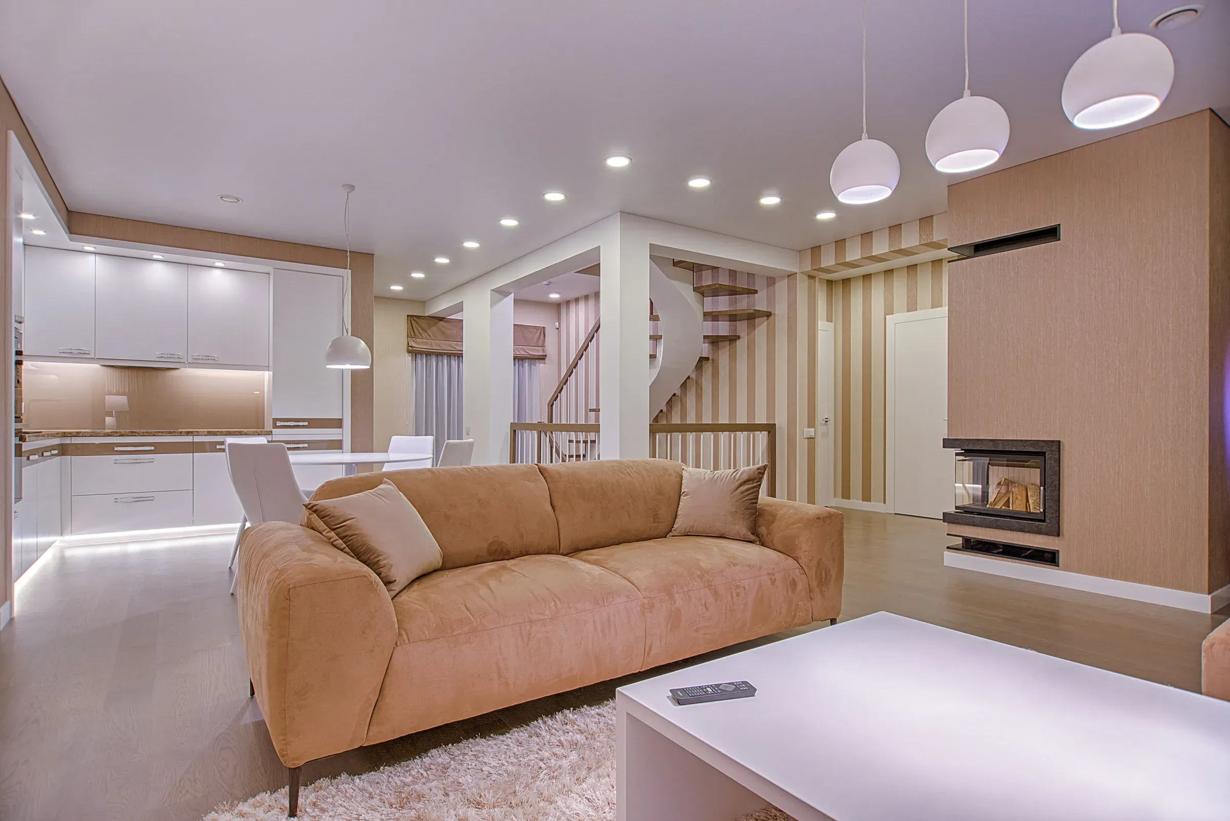 Open-plan living room with a tan sofa, white staircase, fireplace, white pendant lights and kitchen