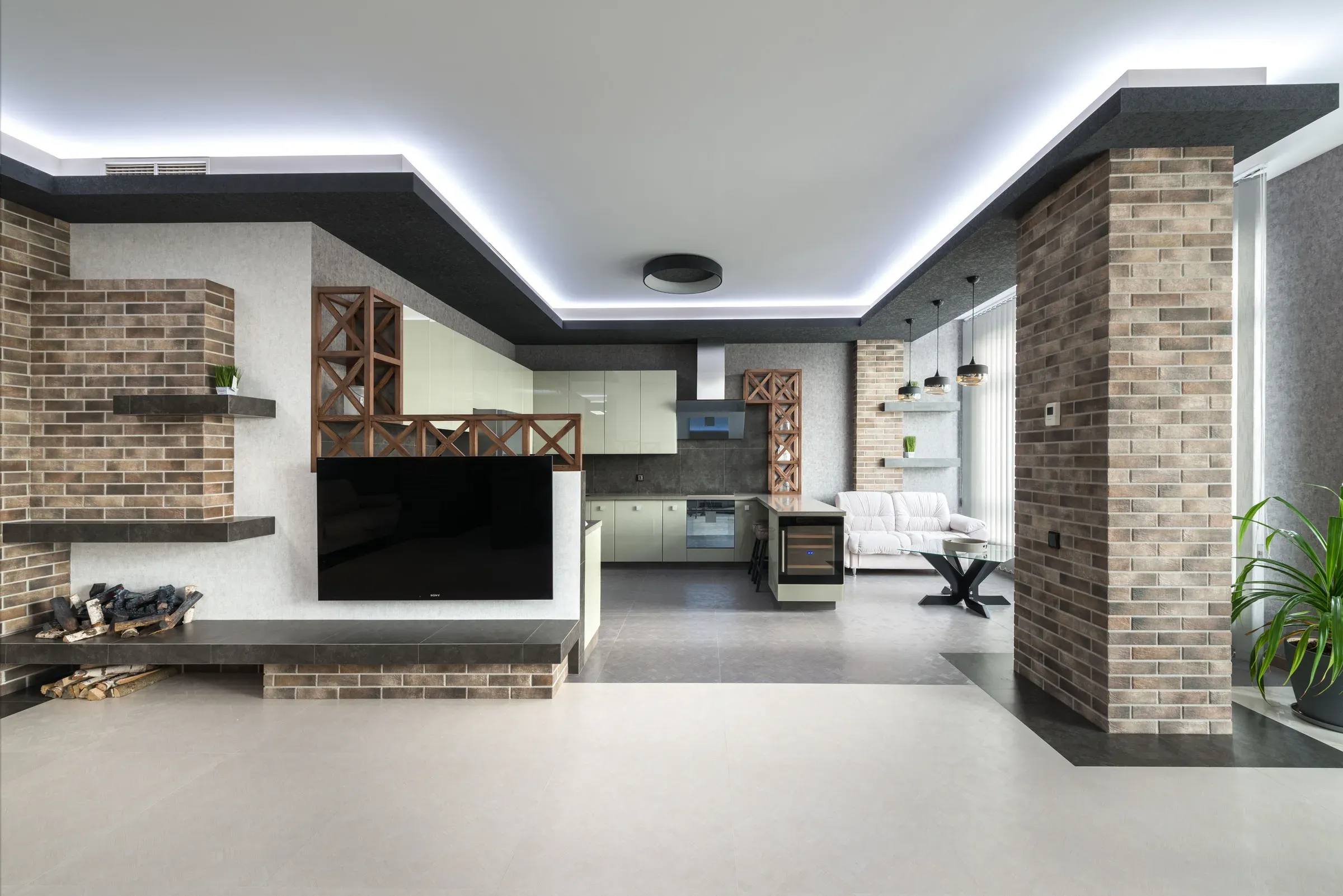 Open-plan living room with a TV on a brick-clad wall, floating shelves, firewood niche and cove lighting
