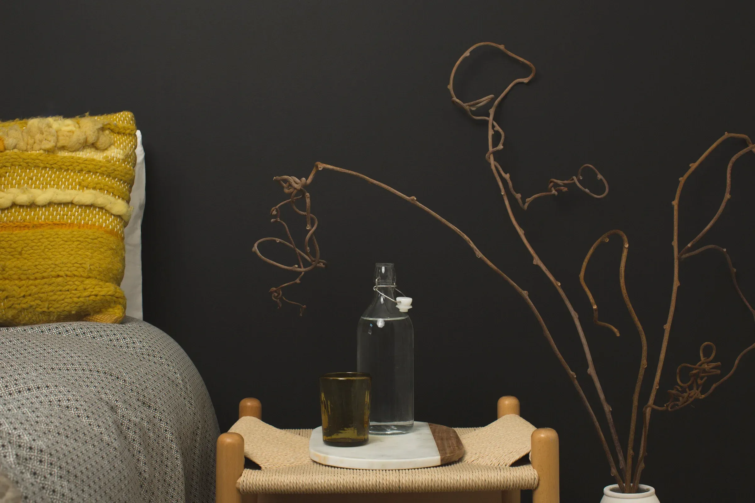 Matte black bedroom wall with a mustard knitted cushion, woven bedside stool and dried branches