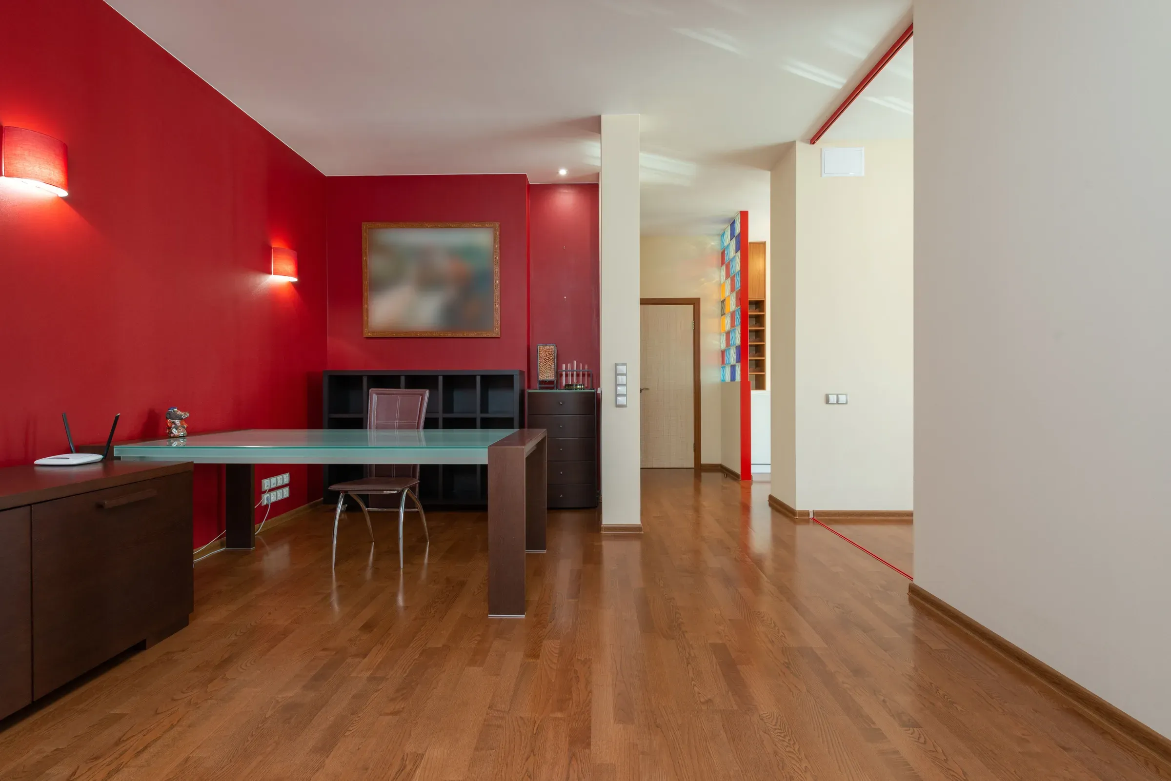 Room with deep red feature walls, wall lights, a glass desk and a warm wooden floor