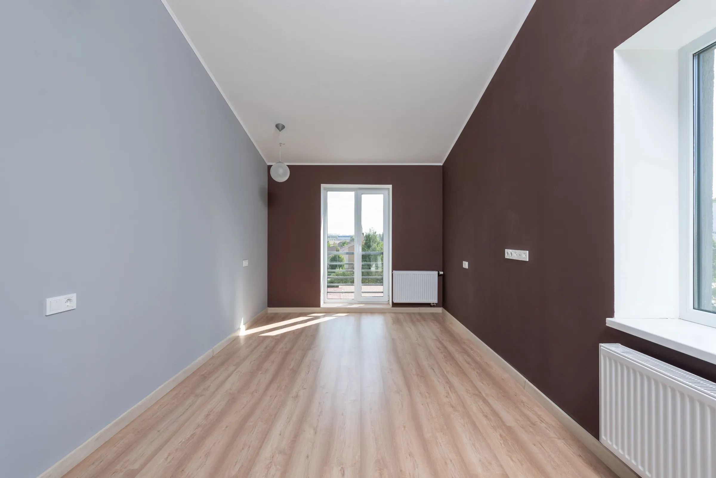 Empty room painted in two colours, grey-blue and chocolate brown, with a light wooden floor and balcony door