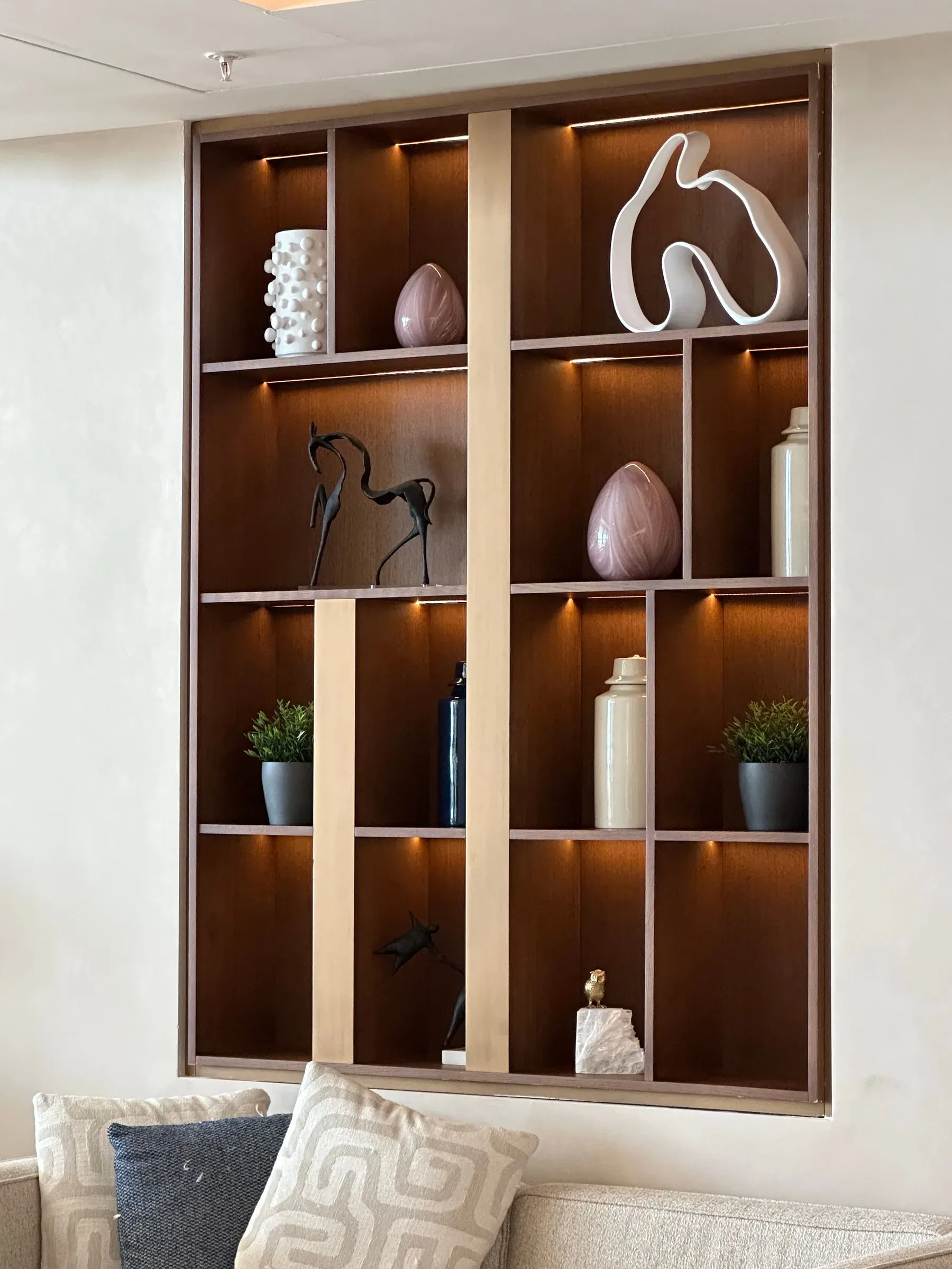 Backlit walnut display niche with sculptures, vases and plants above a sofa