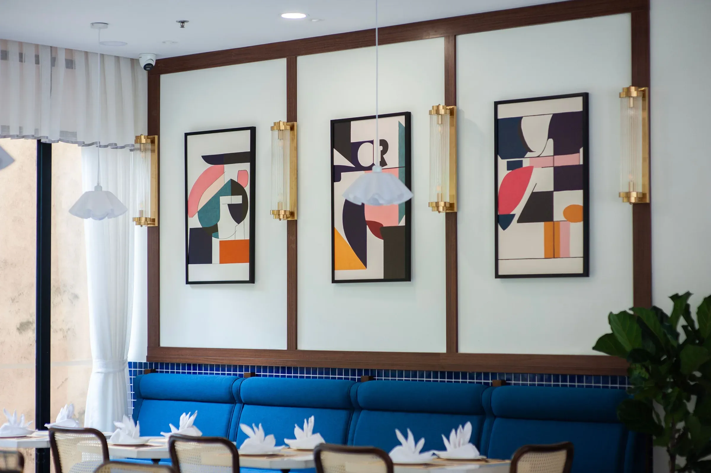 Wall with three framed abstract art prints on white panels between brass sconces above a blue banquette