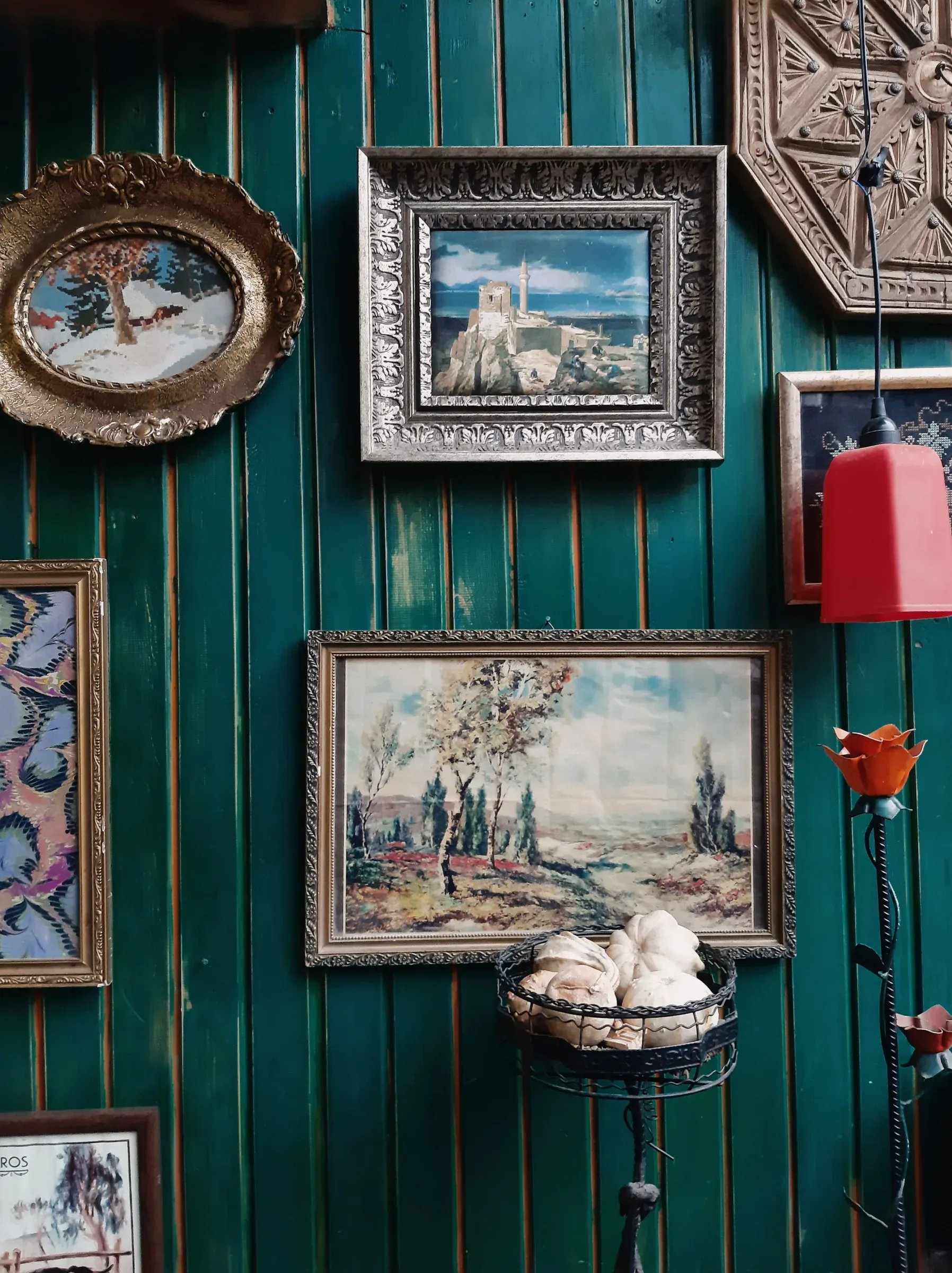Green wood-plank wall hung with vintage framed paintings, an ornate panel and red lamp