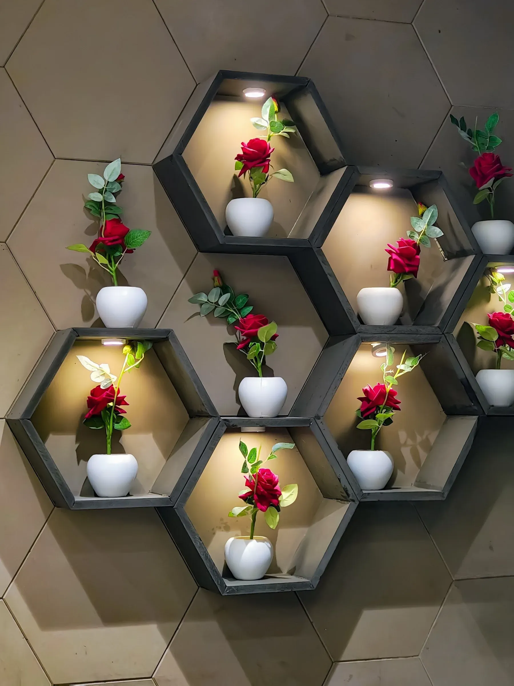 Lit hexagon wall shelves holding white pots of red roses on a hexagon-tile wall