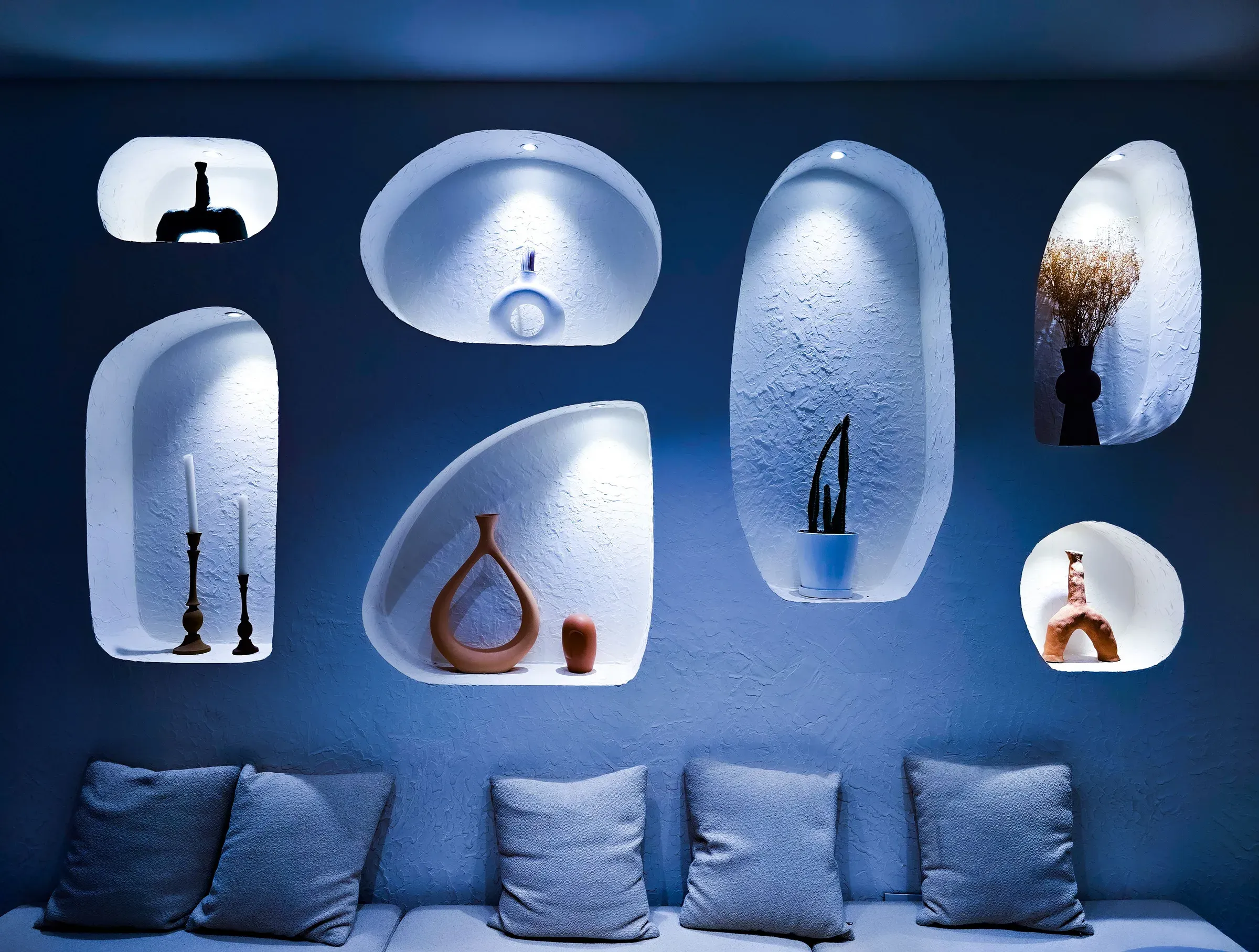 Organic plaster wall niches lit in blue, holding pottery, candles and plants above a cushioned bench