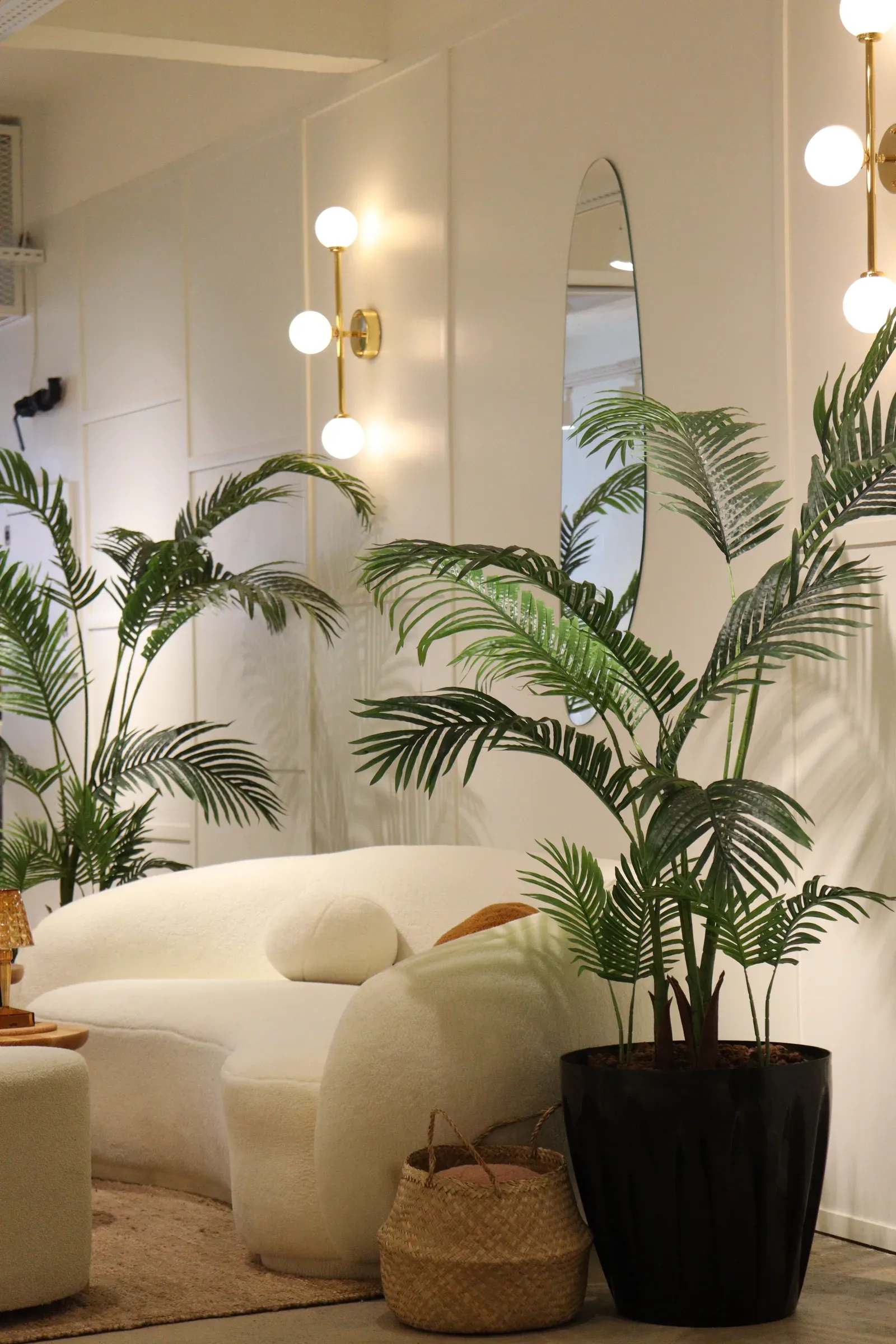 White panelled wall with brass globe sconces, an oval mirror, palms and a boucle sofa