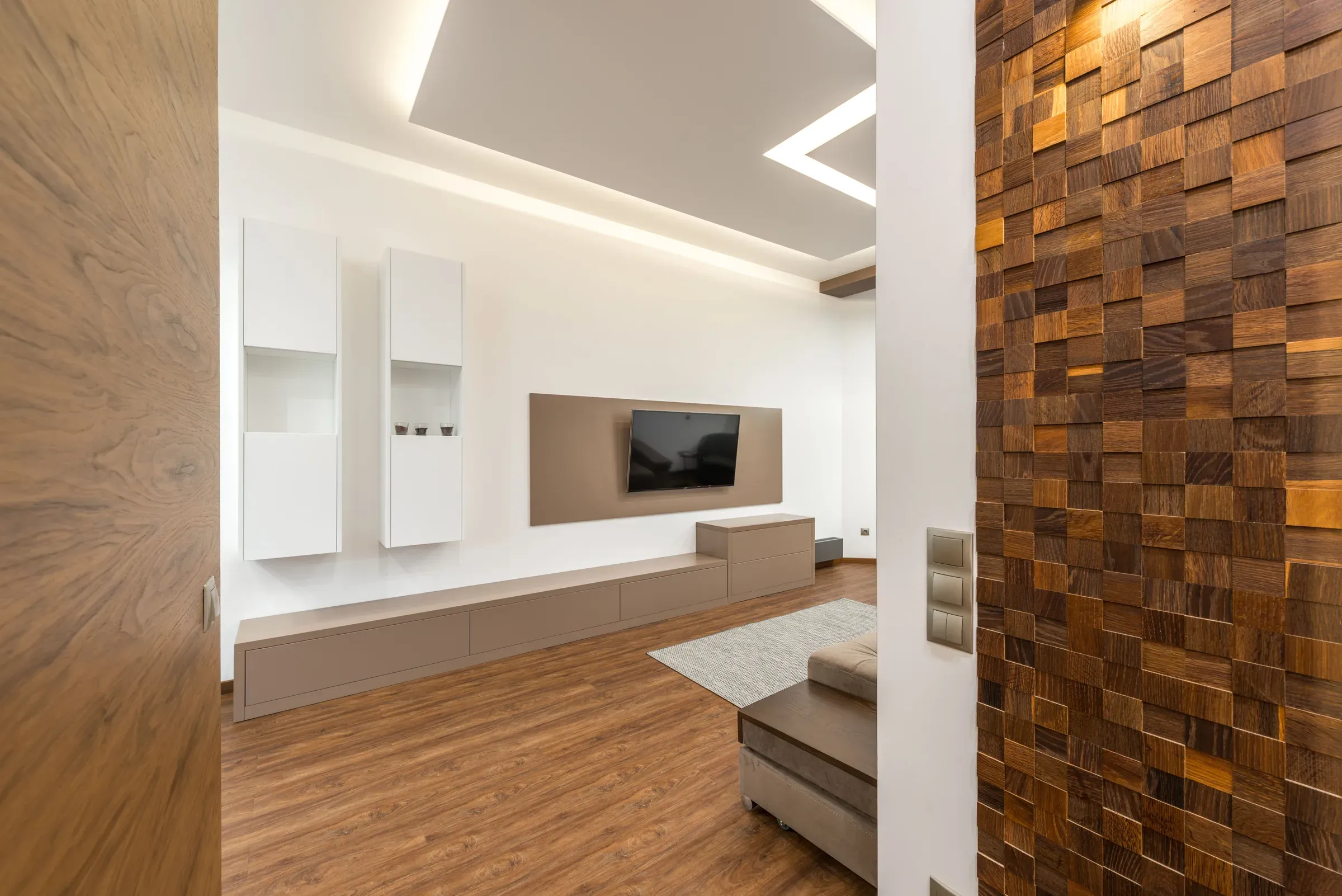 Living room seen past a wooden mosaic cube wall cladding, with a TV panel and floating white cabinets