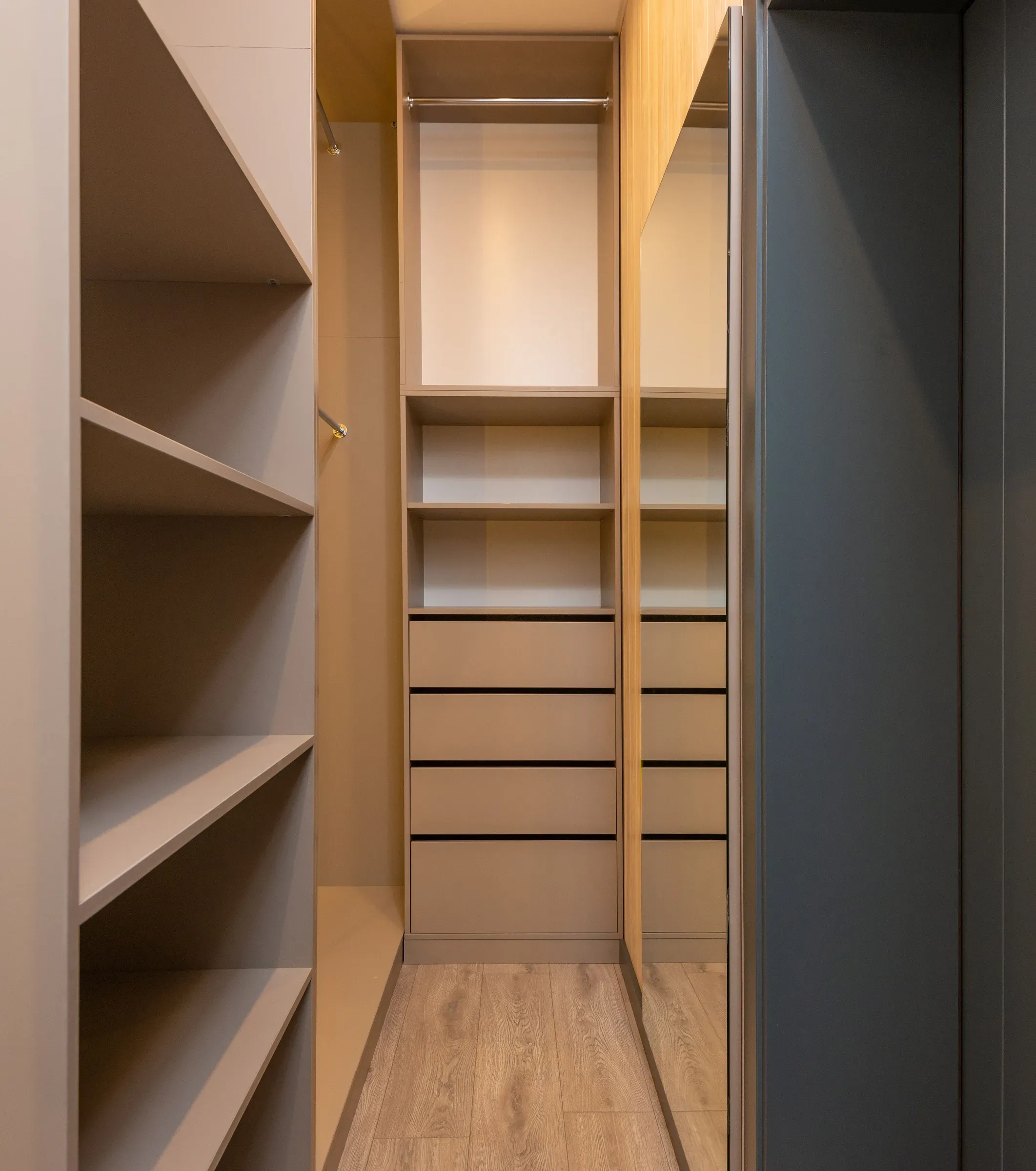 Narrow beige walk-in closet with open shelves, four drawers, hanging rail and a mirrored sliding door