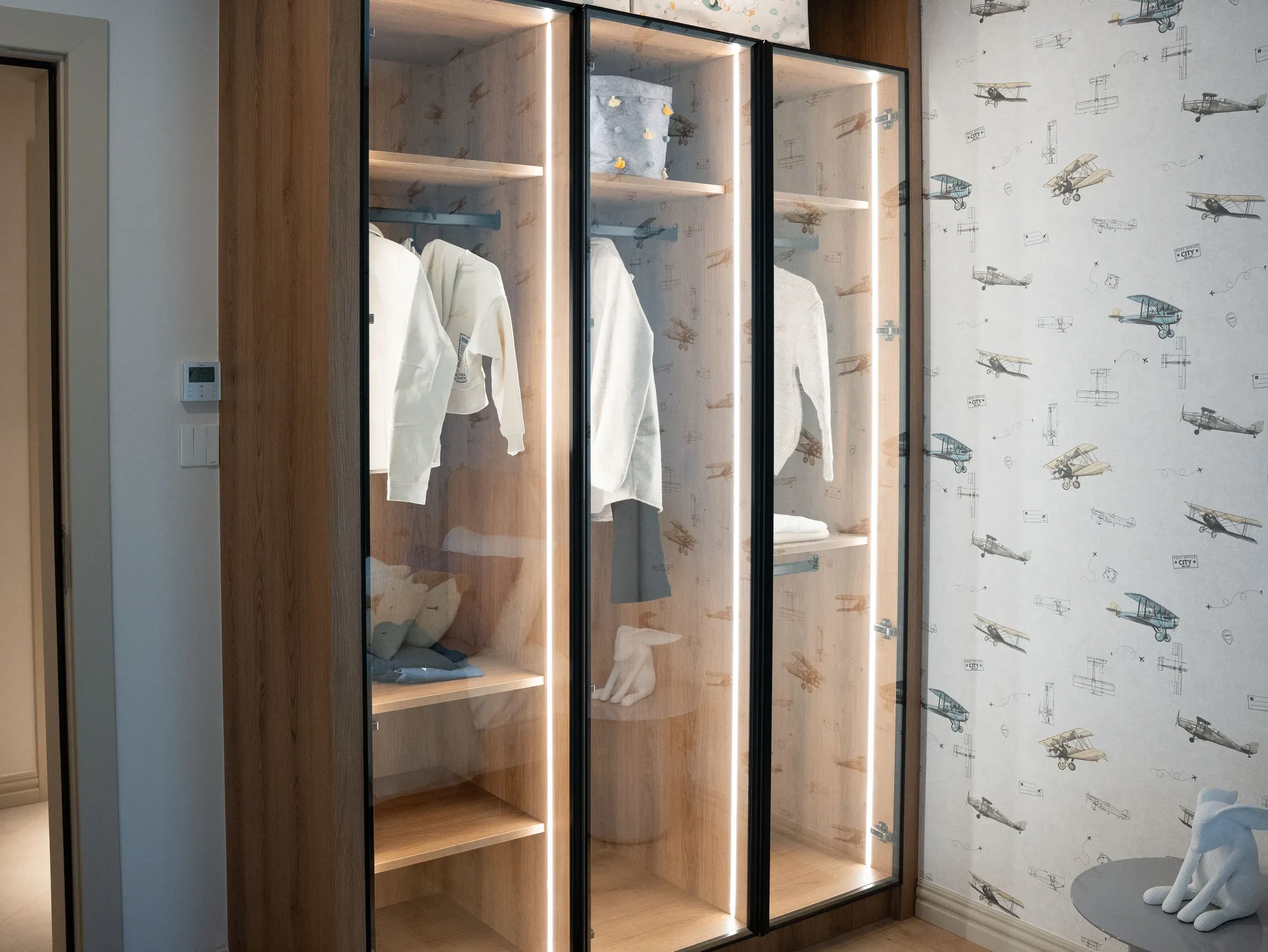 Kids' room wardrobe with glass doors, oak shelves and LED lighting beside airplane-print wallpaper